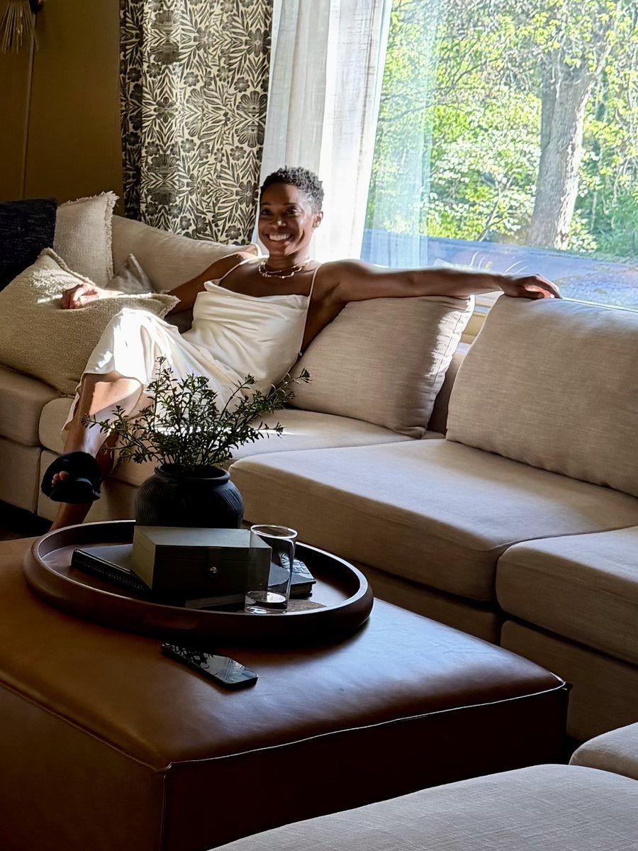 Angela at home in golden window light, laughing, at ease on her couch.